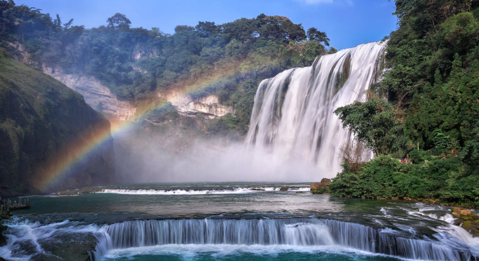 The wide Huangguoshu Waterfall cascading over a lush green cliff, surrounded by dense forest and rising mist.